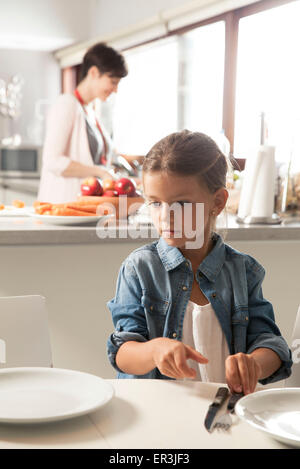 Ragazza impostazione della tabella, madre preparare il cibo in background Foto Stock