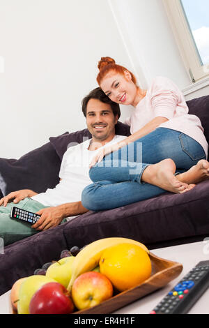 Coppia giovane in appoggio sul divano nel soggiorno mentre l'uomo è di guardare la TV e la donna è la lettura di un libro. Foto Stock