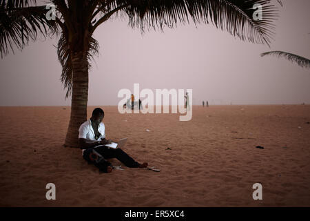 Uomo che utilizza un computer portatile mentre è seduto alla spiaggia sotto una palma, Togbin Plage, Route des Peches, vicino a Cotonou, Benin Foto Stock