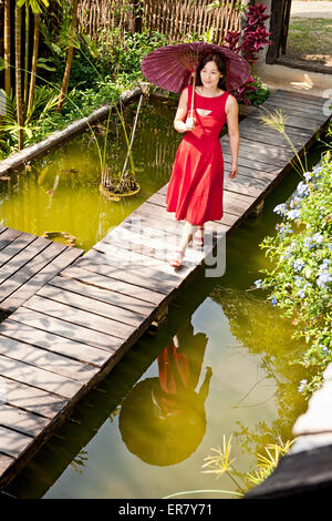 Bella donna in abito rosso a piedi attraverso un giardino Thai Foto Stock