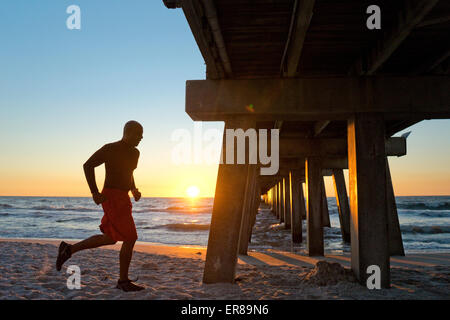 Uomo che corre sotto un molo sulla spiaggia al tramonto Foto Stock