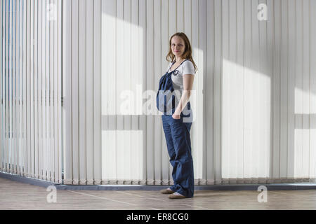 A piena lunghezza ritratto di donna incinta in tuta in piedi contro la tenda Foto Stock