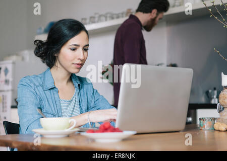 Giovane donna utilizzando laptop con uomo in background in cucina Foto Stock