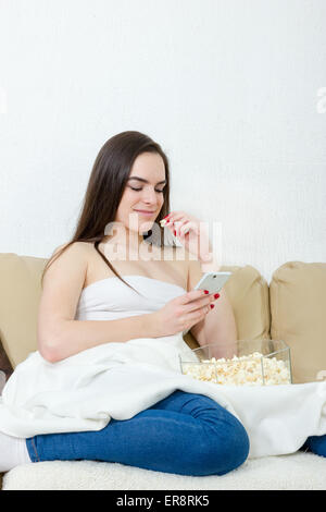 Giovani belle donne digitando messaggio sul telefono cellulare e mangiare popcorn. Da solo felice modello femminile con cellphonel seduta sul letto Foto Stock