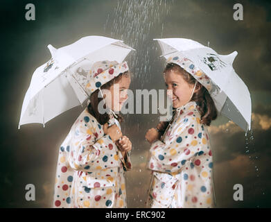 Anni sessanta sorridente TWIN ragazze indossano MATCHING POLKA DOT PIOGGIA DI CAPPOTTI E cappelli in piedi sotto gli ombrelli SOTTO LA PIOGGIA Foto Stock
