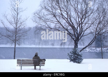 Unico anonimo stagliano uomo seduto da solo sulle fredde WINTERY una panchina nel parco in cerca di neve presso il River Foto Stock