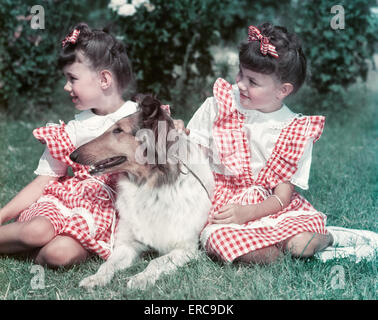 Negli anni quaranta anni cinquanta due gemelle ROSSO DA INDOSSARE ABITI controllato seduta sul prato con COLLIE cane tutto guardando a sinistra Foto Stock