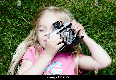 Ritratto di bambina di scattare una foto utilizzando vintage retrò vecchia telecamera cinematografica, giacente su sfondo di erba Foto Stock