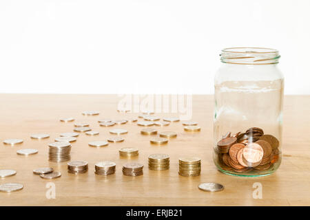 I risparmi in contanti jar. Risparmio di denaro in un vasetto di vetro con il Regno Unito monete impilate e disteso su un tavolo Foto Stock