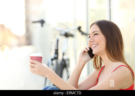 Ragazza chiamando al telefono cellulare e di bere il caffè seduti in un parco con una bicicletta in background Foto Stock