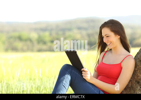 Donna rilassata di lettura di un ebook nel paese appoggiandosi sotto un albero ombra indossando un colore rosso shirt Foto Stock
