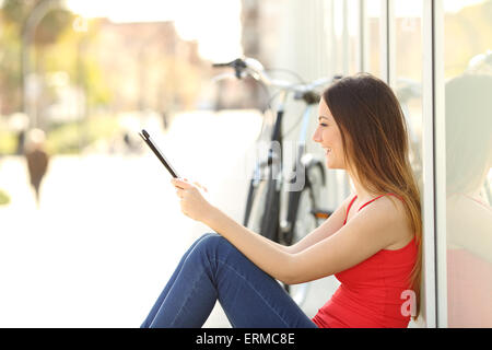 Urban teen ragazza utilizzando un tablet seduta in strada con una bicicletta in background Foto Stock