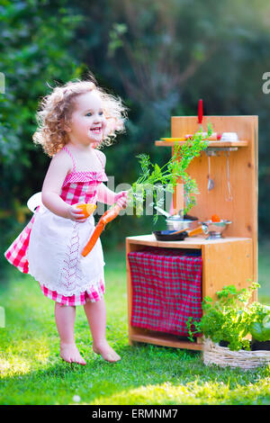 Ricci adorabile ragazza toddler indossando un abito rosso e bianco grembiule di pizzo divertirsi giocando con una bambola giocattolo di cucina nel giardino soleggiato Foto Stock