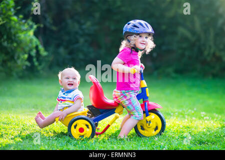 Due bambini felici, ricci adorabile ragazza toddler e una divertente baby boy, fratello e sorella, giocando insieme in sella ad una bici in giardino Foto Stock