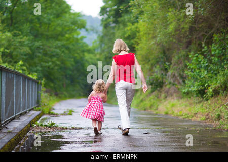 Madre e un po' di parentesi toddler ragazza camminare insieme in un parco sul bagnato estate giorno dopo la pioggia, vista dal retro Foto Stock
