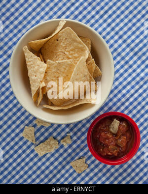 Una ciotola di corn tortilla chips sedersi accanto a un recipiente rosso di salsa su un blu e bianco tovaglia a scacchi. Pezzi di tortilla sono b Foto Stock