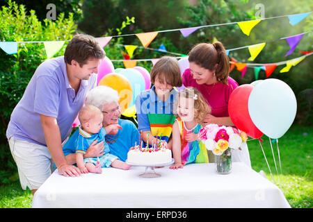 La grande famiglia con tre bambini e la nonna godendo di festa di compleanno con torta soffiando candele in giardino decorato con palloncini Foto Stock