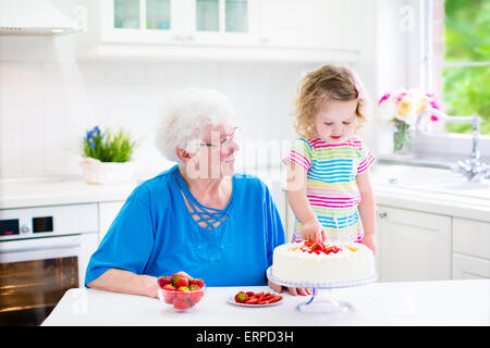 Felice senior lady, amorevole nonna, la cottura a casa la torta di fragole con il suo nipote in un bianco cucina moderna Foto Stock