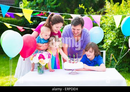 Felice grande famiglia con tre bambini godendo di festa di compleanno con torta soffiando candele in giardino decorato con palloncini e banner Foto Stock