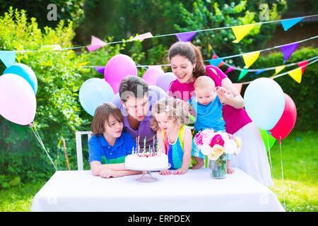 Felice grande famiglia con tre bambini godendo di festa di compleanno con torta soffiando candele in giardino decorato con palloncini e banner Foto Stock