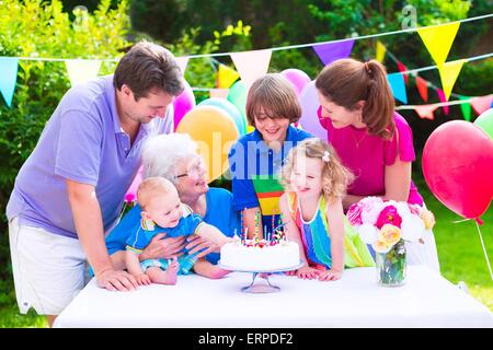 La grande famiglia con tre bambini e la nonna godendo di festa di compleanno con torta soffiando candele in giardino decorato con palloncini Foto Stock