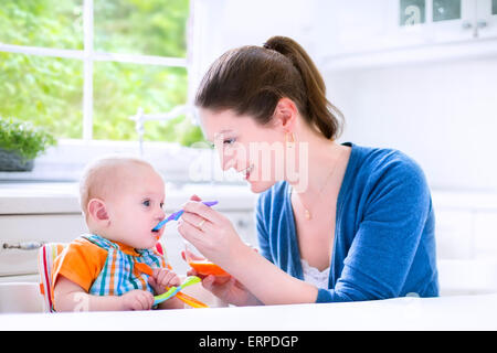 Giovane madre alimentando il suo grazioso baby Figlio conferendogli il suo primo solido cibo sano e puro vegetale dalla carota con un cucchiaio di plastica Foto Stock