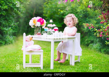 Adorabili bimbi divertenti ragazza con i capelli ricci che indossa un abito colorato per il suo compleanno giocando tea party con un orsacchiotto di peluche bambola Foto Stock