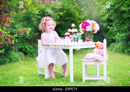 Adorabile ragazza toddler nel giorno del suo compleanno giocando tea party con Teddy bear bambola giocattolo, piatti, tazza di torte e muffin nel giardino soleggiato Foto Stock