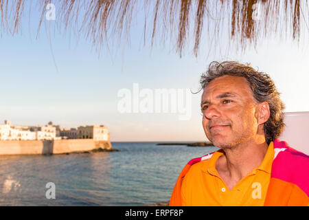 Bello conciate di uomo di mezza età con sale pepe capelli in abbigliamento casual con rosso e arancione maglione giallo e polo shirt in italiano all'aperto: egli è rilassato sotto ombrellone palm mentre antichi edifici sono affacciati sul mare sullo sfondo Foto Stock