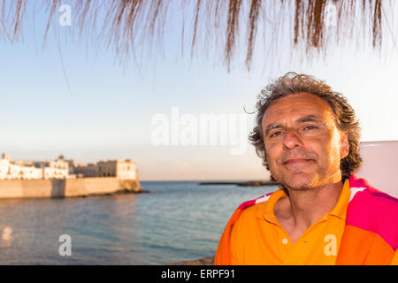 Bello conciate di uomo di mezza età con sale pepe capelli in abbigliamento casual con rosso e arancione maglione giallo e polo shirt in italiano all'aperto: egli è rilassato sotto ombrellone palm mentre antichi edifici sono affacciati sul mare sullo sfondo Foto Stock