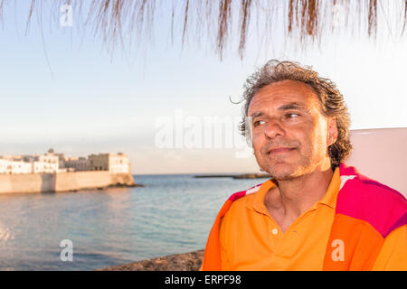 Bello conciate di uomo di mezza età con sale pepe capelli in abbigliamento casual con rosso e arancione maglione giallo e polo shirt in italiano all'aperto: egli è rilassato sotto ombrellone palm mentre antichi edifici sono affacciati sul mare sullo sfondo Foto Stock