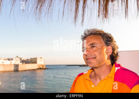 Bello conciate di uomo di mezza età con sale pepe capelli in abbigliamento casual con rosso e arancione maglione giallo e polo shirt in italiano all'aperto: egli è rilassato sotto ombrellone palm mentre antichi edifici sono affacciati sul mare sullo sfondo Foto Stock