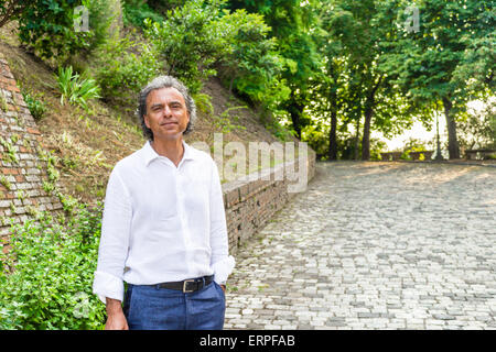 Bello conciate di uomo di mezza età con sale e pepe per i capelli e gli occhi verdi vestito con biancheria camicia bianca e pantaloni blu in medievale italiana all'aperto: egli è rilassato vicino a pareti di mattoni di un edificio medievale Foto Stock