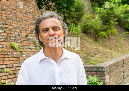 Bello conciate di uomo di mezza età con sale e pepe per i capelli e gli occhi verdi vestito con biancheria camicia bianca e pantaloni blu in medievale italiana all'aperto: egli è rilassato vicino a pareti di mattoni di un edificio medievale Foto Stock