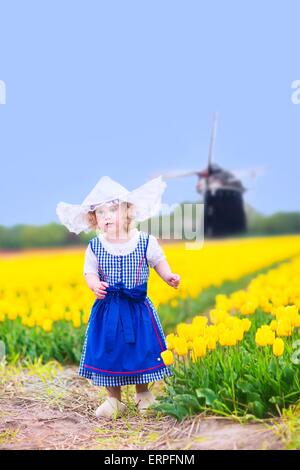 Il Toddler girl indossando Olandese tradizionale costume nazionale il vestito e hat giocando in un campo di tulipani in fiore accanto a un mulino a vento Foto Stock