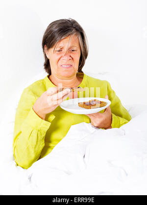 Donna nella sua fifties seduto nel suo letto con una piastra di toast e guardando sconvolto Foto Stock