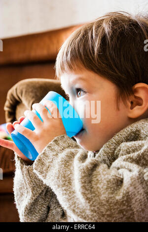 Bambino caucasico, boy, 3-4 anni, seduto su un divano, tenendo in entrambe le mani un bicchiere di plastica e bere da esso mentre si guarda qualcosa nella parte anteriore Foto Stock