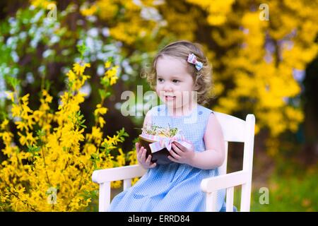 Carino curly toddler ragazza in un abito blu godendo di uovo di Pasqua Caccia nel giardino con giallo fiore fiori di forsitia Foto Stock