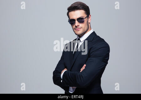 Imprenditore gravi in occhiali da sole in piedi con le braccia incrociate su sfondo grigio Foto Stock