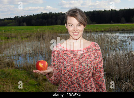 Giovane donna in piedi all'aperto accanto a un lago e tenendo un Apple nella sua mano Foto Stock