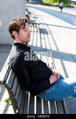 Uomo in camicia nera e jeans seduti su un banco di lavoro con gli occhi chiusi Foto Stock