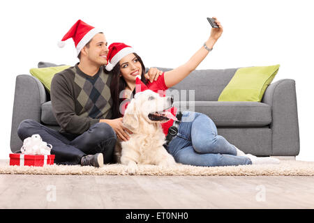 Coppia giovane con cappelli di Babbo Natale prendendo un selfie con il loro cane seduti davanti ad un moderno edificio grigio divano isolati su sfondo bianco Foto Stock