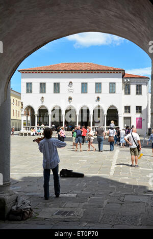 Musicista giocando in ombra di arco al di sotto di Palazzo Pretorio con Tito Square turisti & Palazzo Loggia al di là di Koper Slovenia, Penisola istriana Foto Stock