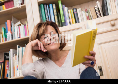 Senior donna seduta di fronte a scaffale e lettura, Monaco di Baviera, Germania Foto Stock
