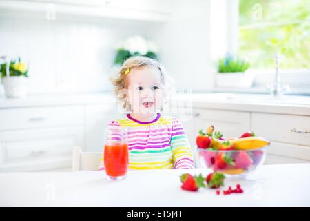 Bellissimo bimbo ragazza con i capelli ricci che indossa una maglietta colorato avente colazione bevendo succo in un bianco cucina soleggiato Foto Stock