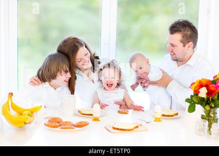 Felice famiglia giovane con tre bambini divertirsi su una Domenica mattina la prima colazione in un bianco sala da pranzo con un grande finestra Foto Stock