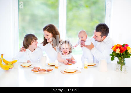 Felice famiglia giovane con tre bambini divertirsi su una Domenica mattina la prima colazione in un bianco sala da pranzo con un grande finestra Foto Stock