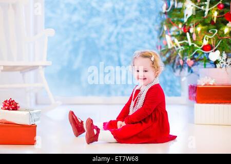 Carino curly piccola ragazza in un abito rosso e bianco perla collana giocare sotto un albero di Natale con presenta seduta sul pavimento Foto Stock