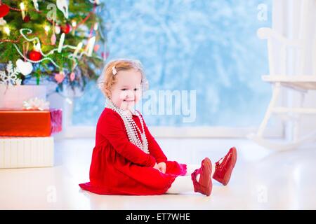 Carino curly piccola ragazza in un abito rosso e bianco perla collana giocare sotto un albero di Natale con presenta seduta sul pavimento Foto Stock