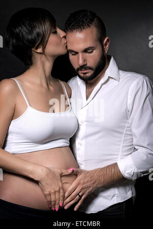 Dolce matura in attesa di baby, bella donna incinta kiss il marito su sfondo nero, tenendo le mani sulla pancia Foto Stock